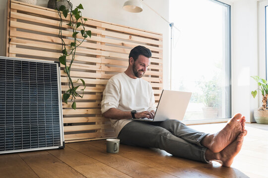 Happy businessman sitting by solar panel and using laptop at home