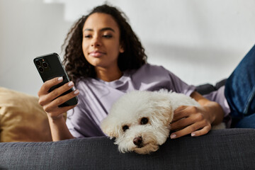 A woman relaxes on a couch, holding her bichon frise dog and a cell phone.