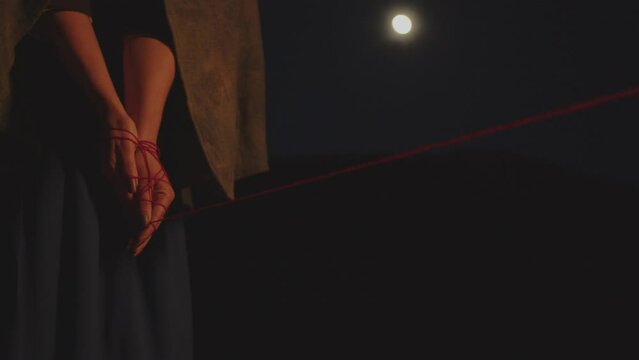 A woman in vintage clothing in the dark with her hands tied with red thread against the backdrop of the full moon. Soft light on hands. Psychology