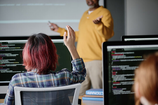 Back view shot of unrecognizable teen girl sitting at desk in Computer Science classroom raising her hand during lesson