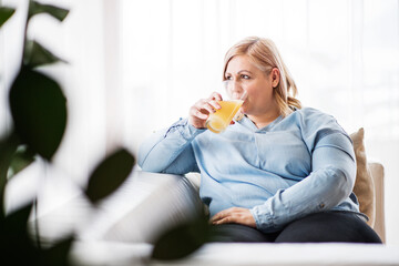 Portrait of beautiful young overweight woman at home, drinking orange juice, enjoying sweet taste.