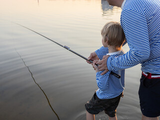 Dad teaches his son to fish with a fishing rod, relaxing and fishing together at sunset on the lake. father's day concept. favorite hobby of parents and children. be like dad, teach your son to fish