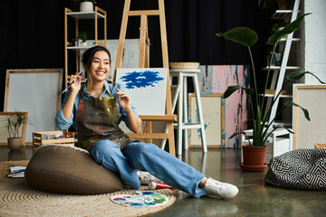 A young Asian artist takes a break from painting, sitting on a beanbag in her art-filled studio.