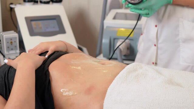 Cosmetologist doctor chooses program and begins cavitation procedure in abdominal area in fight against fat and cellulite in beauty salon. Woman getting anti fat therapy on stomach, close up. Hardware