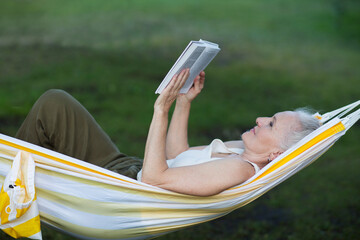 Slow life. Enjoying the little things. lifestyle portrait senior woman with gray hair rests and reads a book lying in a hammock in a summer park or garden.