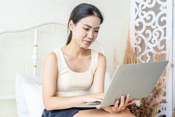 Fototapeta premium Relaxed beautiful young asian woman with sheet facial mask lying working on laptop computer on bed, beauty girl applying face mask for skincare of wrinkle at home, skin care, health and wellness.