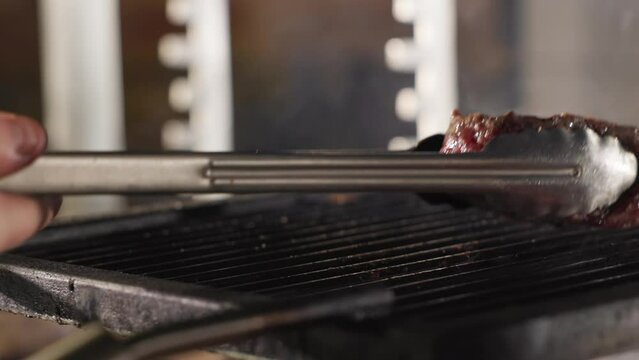 Chef hand flipping piece of meat with grill marks with metal tongs while it cooked on hot grate. Concept of processing of ingredients, preparing and restaurant.