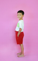 Portrait of smiling Asian boy ages 7 years old put hand in his pocket of pant on pink studio background. Image body full length side view.