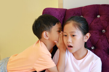 Close-up Asian little boy kid whispering a something secret to young girl child ear. Brother and sister communication concept.