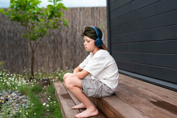 Cute pre-teen boy in headphones listening to music on a wooden terrace outdoors
