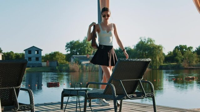 Stunning brunette in a black mini skirt, white tank top, smart watch, and sunglasses comes to sunbed on a luxury resort terrace with a breathtaking river view. She put handbag beside her and lays down