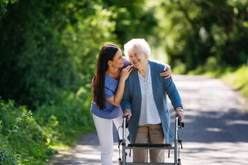 Female caregiver and senior woman with walker on walk in nature. Nurse and elderly woman enjoying a...