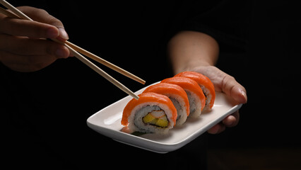 Hands holding plate of Uramaki sushi or California salmon roll on black background. Japanese food style concept