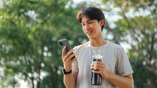 Happy asian man holding mobile phone and bottle of water at public park, Male using smartphone, checking mobile fitness application, copy space.