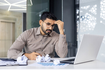 Frustrated businessman sitting at desk with crumpled papers, analyzing documents on laptop in modern office environment.