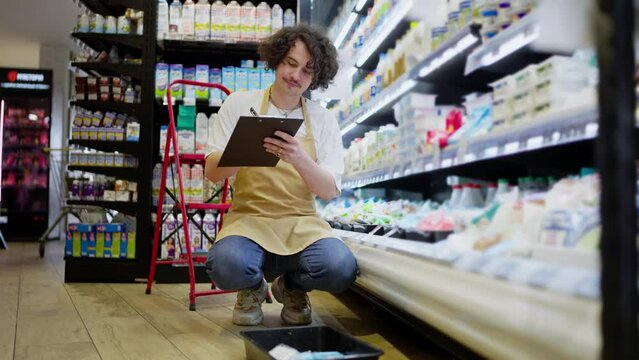 A guy with curly hair with a tablet in his hands takes inventory of goods in the dairy products compartment in a supermarket