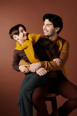 A father and son share a heartwarming moment, both smiling brightly while sitting on a stool in front of a brown background.