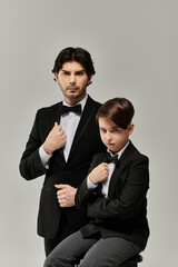 A father and son pose in matching tuxedos, the son sitting, the father standing.