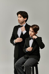 A father and his son stand side by side, both dressed in black suits and bow ties, ready for a formal event.