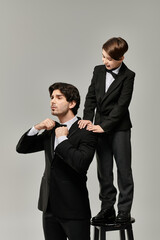 A father adjusts his bow tie while his son stands on a stool and looks on.