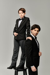 A father and son, both in black suits, stand together in a studio setting.