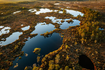 An aerial view of wetlands during sunset, featuring reflective water bodies interspersed with patches of land and vegetation. The sun's rays cast a golden glow over the serene landscape