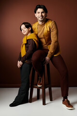 A father and son pose for a portrait, the father sitting on a stool and the son standing beside him. Both are wearing brown and yellow clothing.