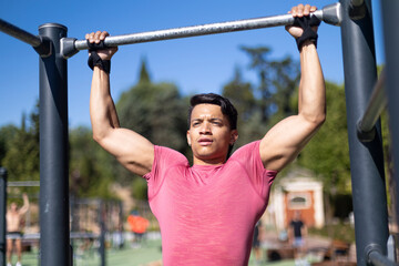 Latin man in pink shirt performing calisthenics pull-ups on outdoor gym equipment in park.