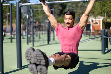 Young latin man in pink shirt doing leg raises on outdoor calisthenics equipment.