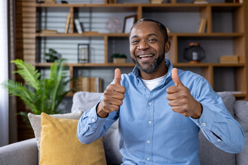 Cheerful man smiling and giving a thumbs up gesture while sitting on a sofa in a comfortable home environment ,with shelves and plants in the background.