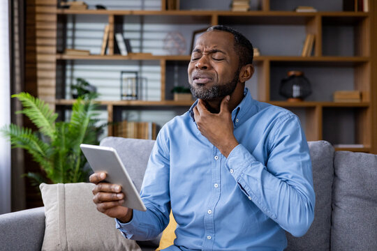 Man experiencing throat pain while consulting a doctor online via video call in a home office setting. Holding a tablet and displaying discomfort.