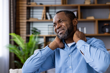 A man is sitting indoors, visibly suffering from neck pain. He is holding his neck with a pained expression, indicating discomfort.