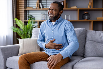 Man clutching his stomach and expressing discomfort while sitting on a sofa in a modern living room. Potential scene related to health issues or digestive problems.