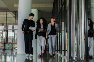 Three people are walking down a hallway, one of them holding a piece of paper. Scene is professional and serious, as the people are dressed in business attire