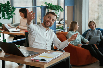 Cheerful friendly man is sitting by the table with laptop. People are working in the office with bean bags chairs in it