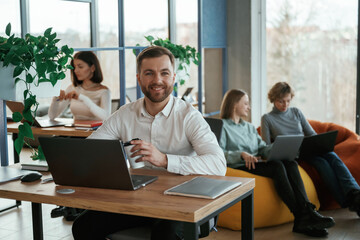 Cheerful friendly man is sitting by the table with laptop. People are working in the office with bean bags chairs in it