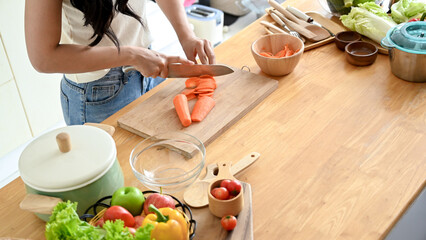 A close-up image of a woman slicing carrots on a cutting board at a wooden kitchen tabletop, cooking in the kitchen. healthy food, lifestyle, home cooking