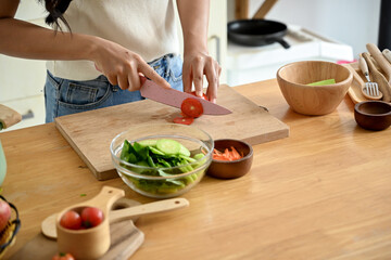 A beautiful, happy young Asian woman cutting a tomato on a cutting board, preparing ingredients for her healthy salad, enjoying cooking in the kitchen. home cooking, domestic life, healthy eating