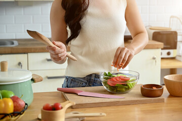 An attractive Asian woman is in the kitchen, holding a bowl of salad and a wooden spatula, enjoying...