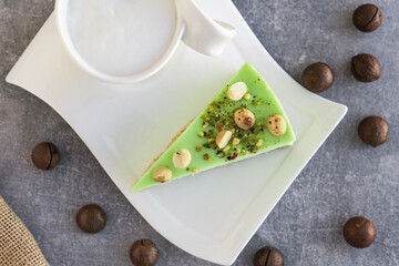 Green pistachio cake with macadamia nuts on white plate with cappuccino. Creamy pistachio cheesecake