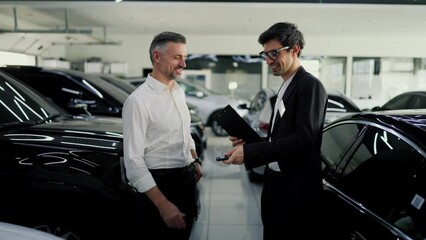 Happy middle-aged man with gray hair in a white shirt shakes hands with an assistant in a business suit while purchasing his new car at a car dealership