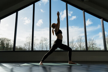 Woman is practicing yoga vinyasa flow, warrior asana pose yoga exercise indoors, against the window. Wellness, lifestyle, copy space.