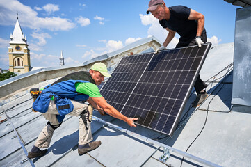 Workers building solar panel system on metal rooftop in urban setting. Two men installers carrying...