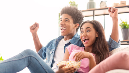 A teen couple is sitting on a couch in their living room, excitedly watching television while eating popcorn. Both have happy expressions on their faces, suggesting they are enjoying the show.