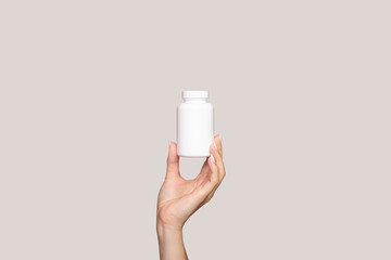 A young woman's hand holds a white plastic bottle isolated on a beige background. Mockup for...