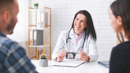 A lady doctor is sitting at a desk in her office, smiling and pointing at an ultrasound image of a baby. A man and a woman are sitting across from her.