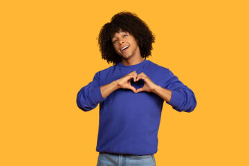 This image shows a young man with an curly hairstyle smiling and making a heart shape with his...