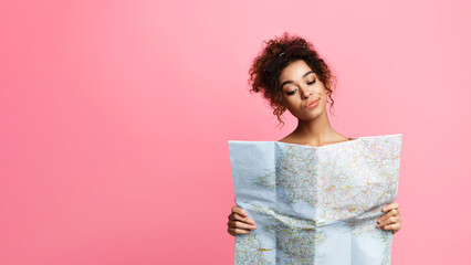 Fototapeta premium A young African American woman with curly hair stands against a soft pink backdrop, carefully examining a large unfolded map. She appears to be planning a journey, copy space