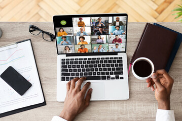 A man sits at a desk with a laptop open, attending a video conference meeting with several other people. The laptop screen displays a grid of video feeds of the participants, view above