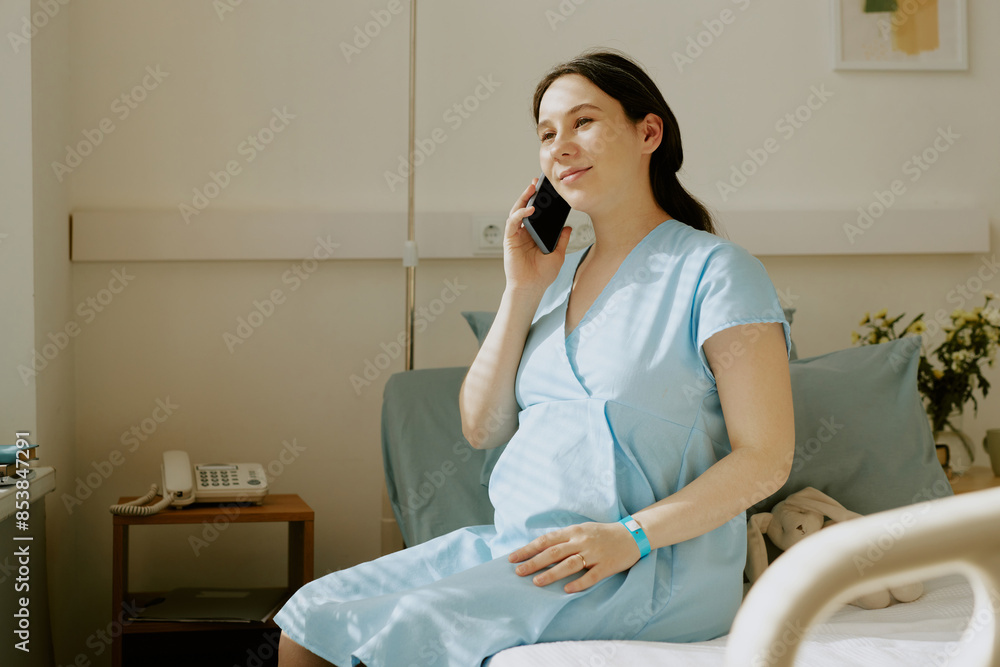 Wall mural Pregnant female patient sitting on bed while she making call and smiling lightly in hospital ward